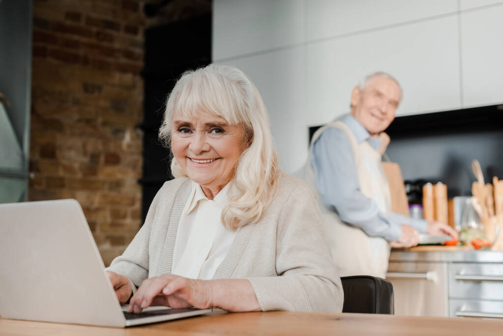 sorridente moglie anziana utilizzando il computer portatile mentre il marito cucina in cucina durante la quarantena
 - Foto, immagini