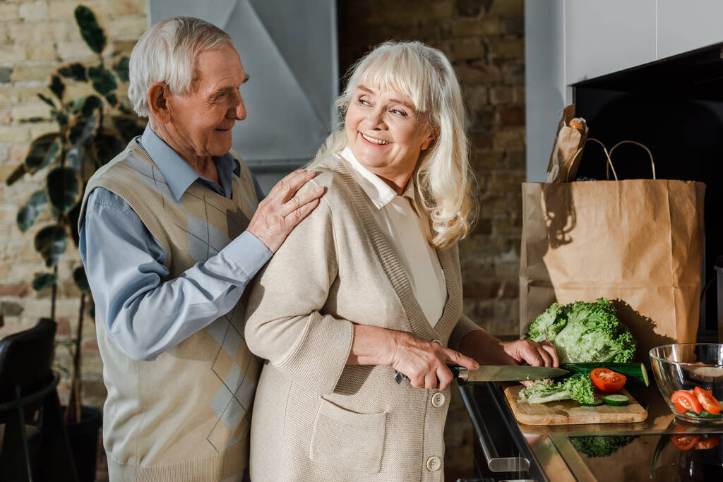 uśmiechnięta para w podeszłym wieku gotująca razem w kuchni podczas samoizolacji  - Zdjęcie, obraz