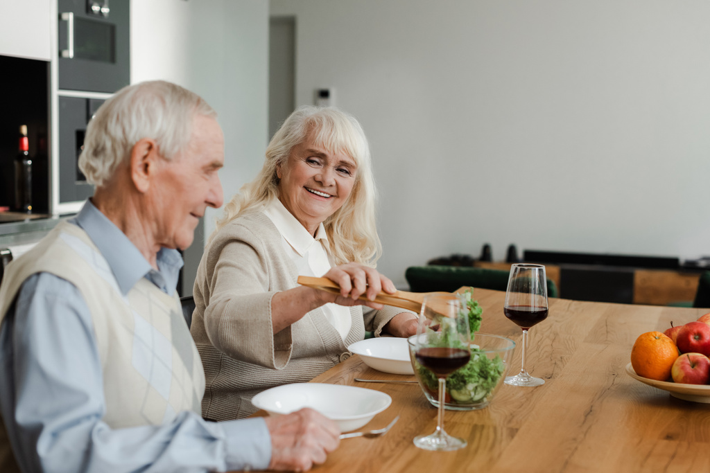 sonriente pareja de ancianos cenando con vino y ensalada en casa en auto aislamiento
 - Foto, Imagen