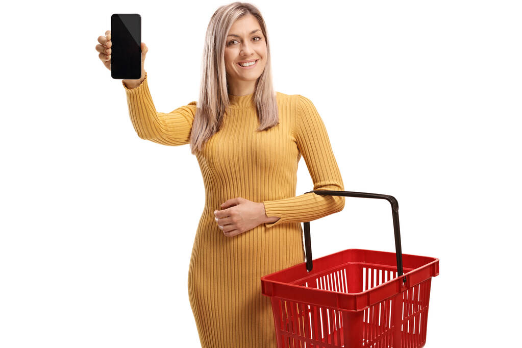 Žena drží prázdný nákupní košík a ukazuje smartphone izolované na bílém pozadí - Fotografie, Obrázek