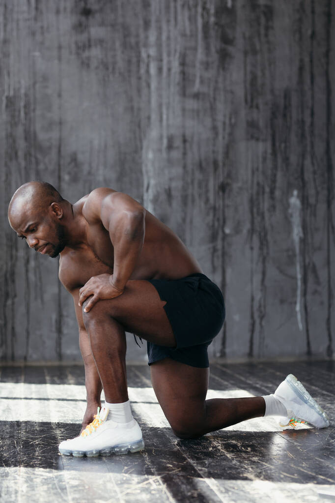 Un atleta negro se para sobre una rodilla, demuestra músculos. Desnudo afroamericano en pantalones cortos negros
 - Foto, Imagen
