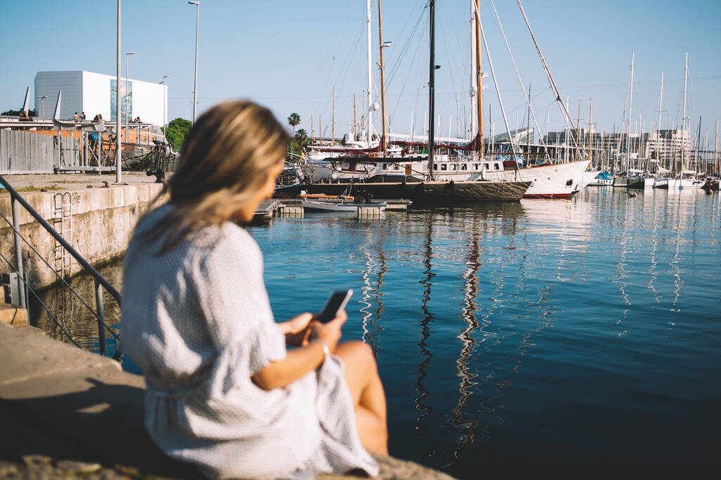 Rückansicht des Millennial-Hipster-Mädchens, das sich im Hafen mit Segelbooten ausruht, mit Smartphone-Gadget für Suchinformationen und Website mit Online-Reiseführer, Frau auf verschwommener Front, die in der Nähe des Meeres sitzt - Foto, Bild
