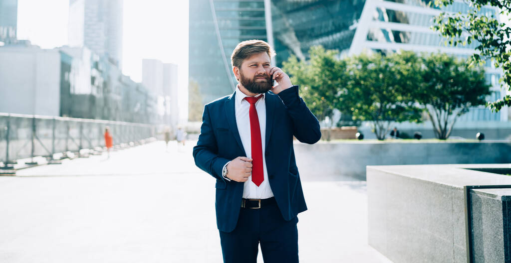 Nachdenklicher bärtiger Mann im dunkeleleganten Anzug und roter Krawatte hört aufmerksam zu, während er auf dem Platz des Business Centers mit dem Handy spricht - Foto, Bild