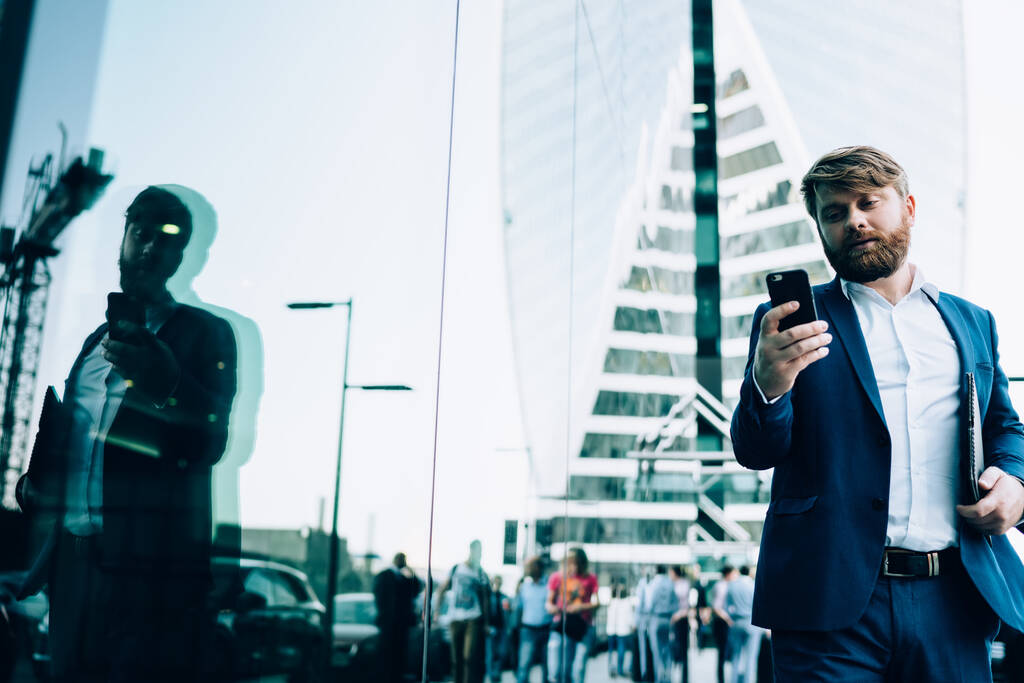 From below calm adult man in elegant blue suit and white shirt with papers focusing on screen of smartphone while standing on street against blurred modern high rise building with mirror facade in downtown - Photo, Image