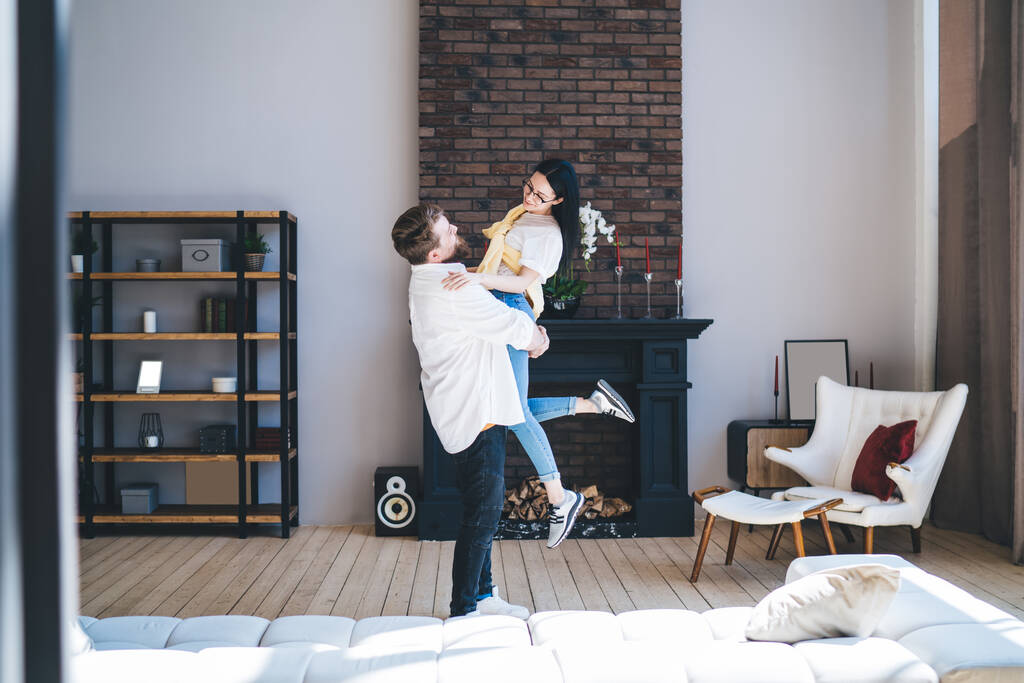 Side näkymä mies kuljettaa rakastava nainen käsissä katsellen toisiaan, kun seisoo tilava moderni olohuone uudessa asunnossa - Valokuva, kuva
