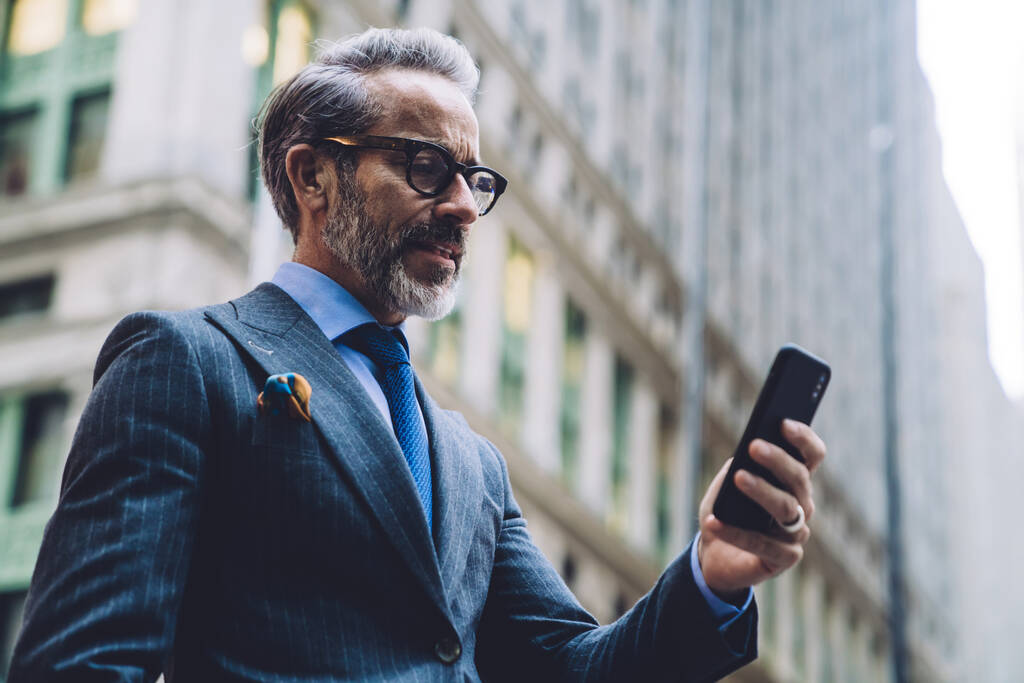 Desde abajo vista lateral del hombre de negocios gris de mediana edad en gafas y elegante traje de negocios oscuro con teléfono inteligente en el fondo del edificio alto en Nueva York
 - Foto, Imagen