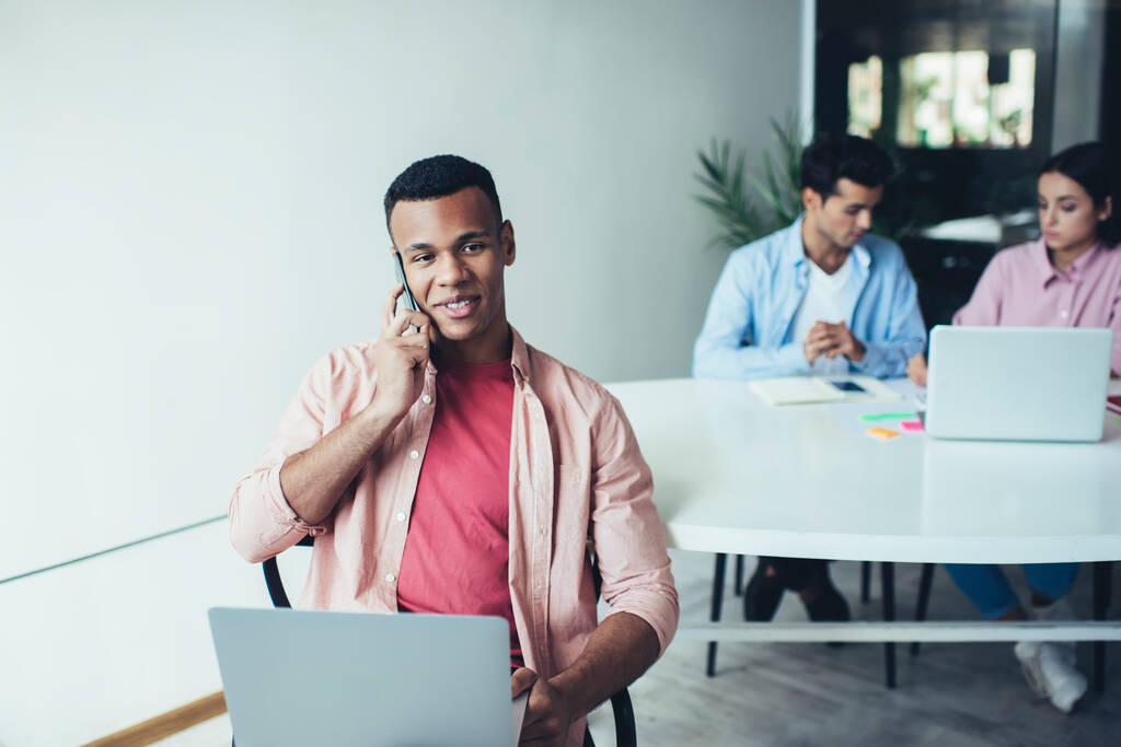 Щасливий чорний чоловік у повсякденному вбранні посміхається і розмовляє по смартфону, сидячи біля столу з колегами на сучасному робочому місці
 - Фото, зображення