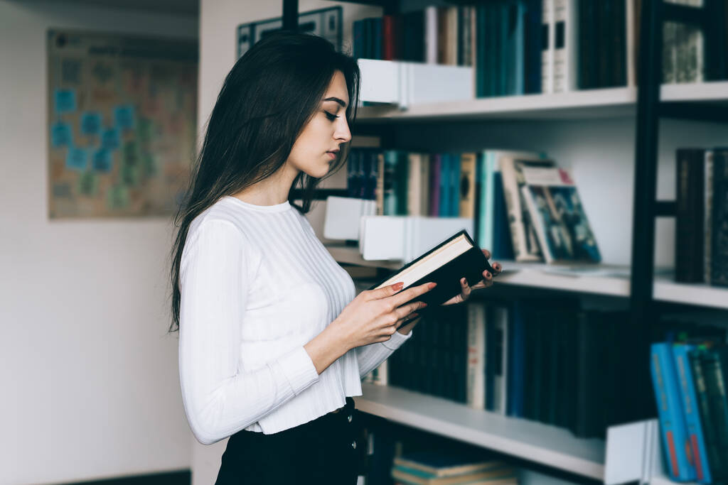 Ευφυής έξυπνη νεαρή γυναίκα με μακριά σκούρα μαλλιά σε λευκό πουλόβερ ανάγνωση βιβλίο στέκεται απέναντι από τη βιβλιοθήκη στη βιβλιοθήκη της πόλης - Φωτογραφία, εικόνα