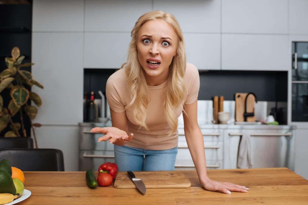 rozzlobená žena gestikuluje a dívá se na kameru v blízkosti čerstvé zeleniny v kuchyni - Fotografie, Obrázek