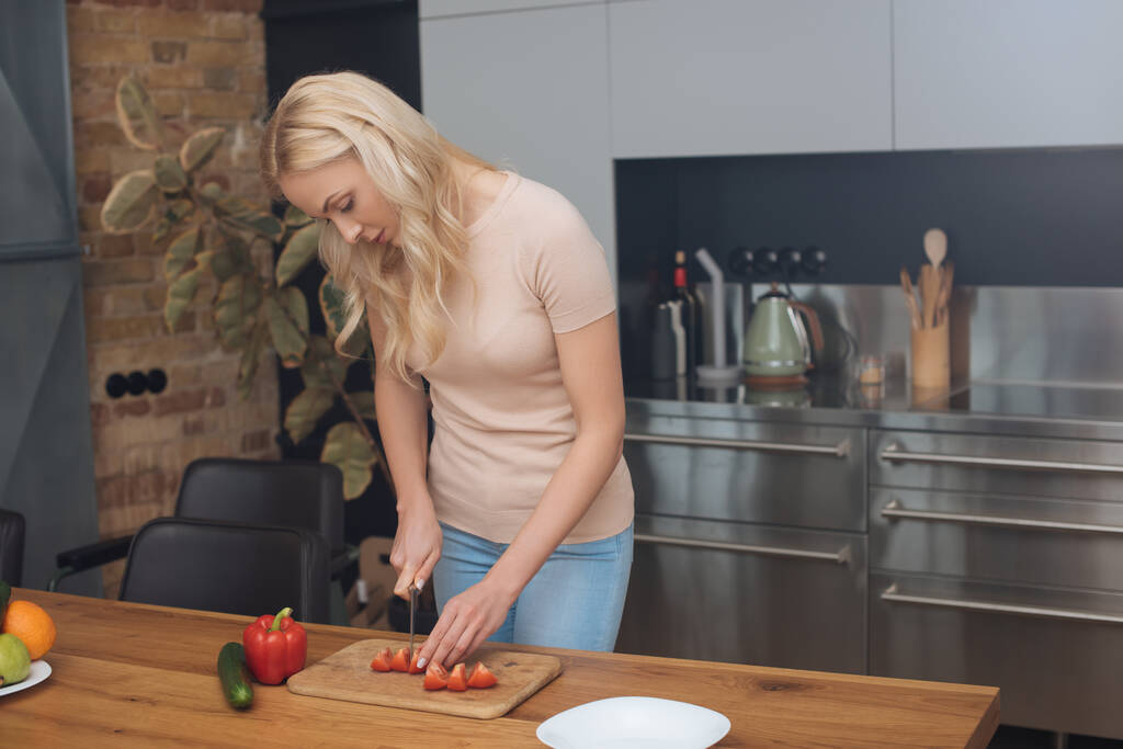 jong blond vrouw snijden verse groenten tijdens het koken in moderne keuken - Foto, afbeelding