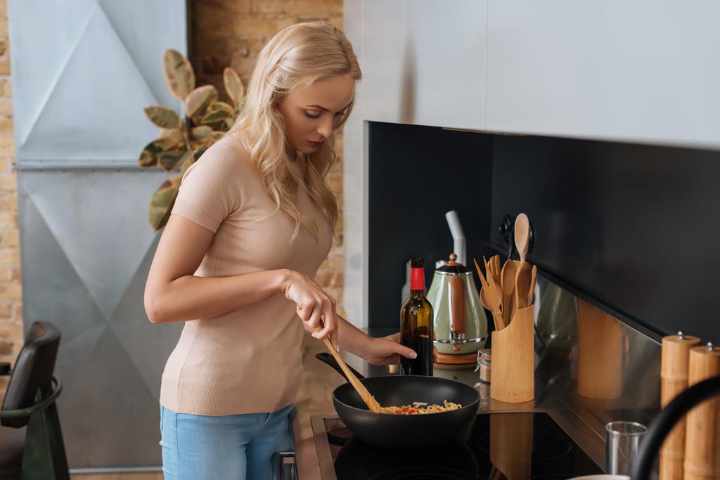 young blonde woman holding bottle of red wine while preparing thai noodles in wok - Photo, Image