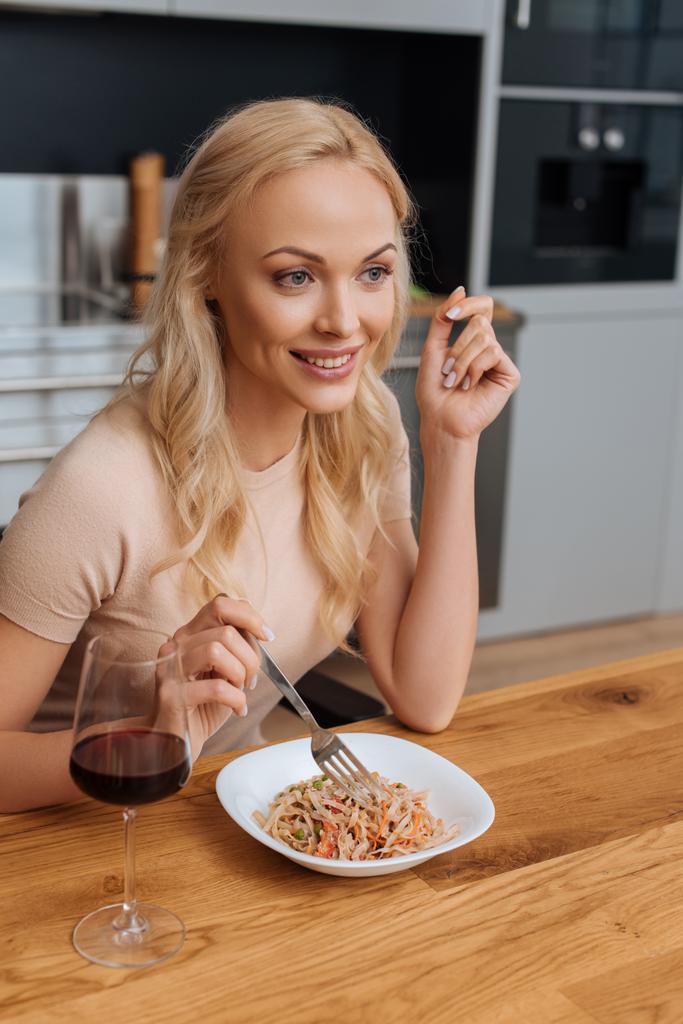 mujer alegre sosteniendo tenedor cerca del plato con fideos tailandeses y copa de vino tinto
 - Foto, Imagen