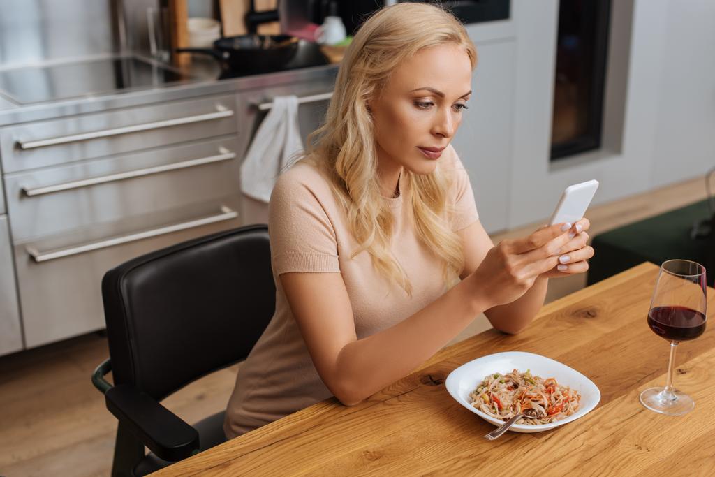 giovane donna che chatta sullo smartphone mentre siede vicino al piatto con tagliatelle tailandesi e bicchiere di vino rosso
 - Foto, immagini