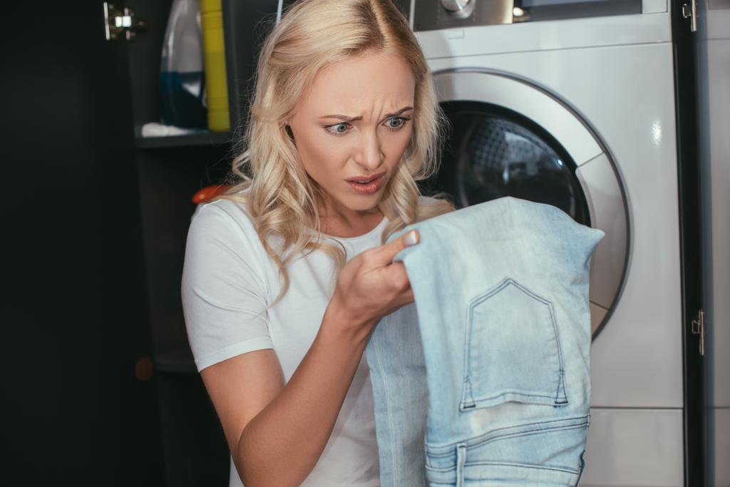 Ev hanımı çamaşır makinesinin yanında dururken kot pantolona bakıyor. - Fotoğraf, Görsel