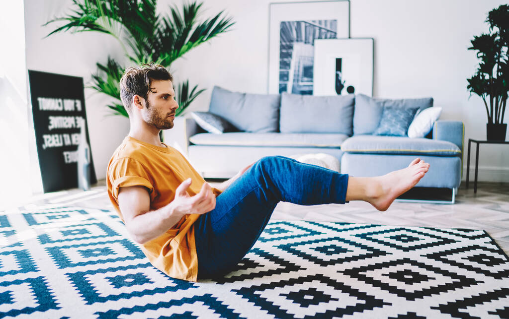 Widok z boku elastyczny mężczyzna w czerwonej koszuli i niebieskie spodnie robi jogę asana siedzi na dywanie w pokoju z sofą w tle - Zdjęcie, obraz