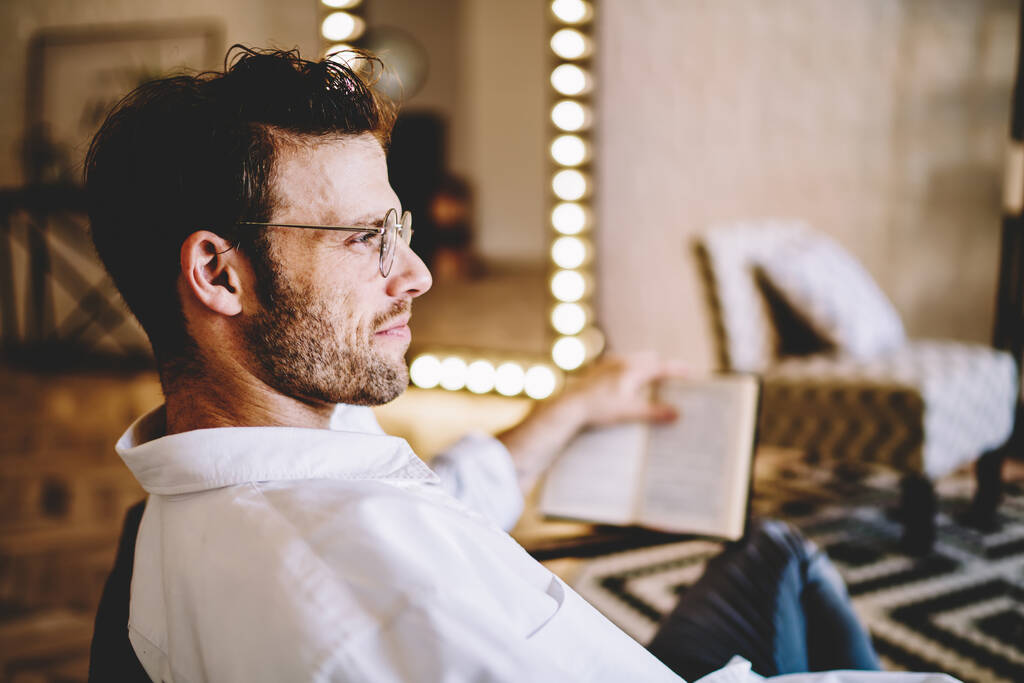 Boční pohled na mladého vousatého muže v brýlích v bílé košili a bílé tričko čtení knihy sedí v křesle v pokoji s černou a bílou carpe - Fotografie, Obrázek