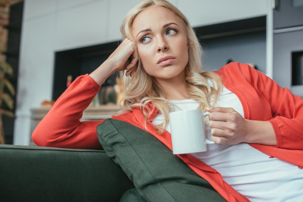 mujer triste mirando hacia otro lado mientras está sentada en el sofá con una taza de té
 - Foto, Imagen