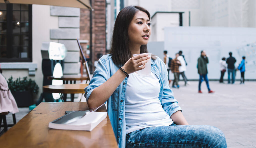 Chilling fêmea étnica ter vidro de bebida refrescante frio e olhando para longe enquanto sentado no café ao ar livre em mesa de madeira com livro
 - Foto, Imagem