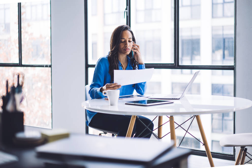 Foco seletivo da mulher afro-americana de sucesso usando laptop e se comunicando no smartphone na mesa redonda branca e estudando contrato no escritório com interior minimalista
 - Foto, Imagem