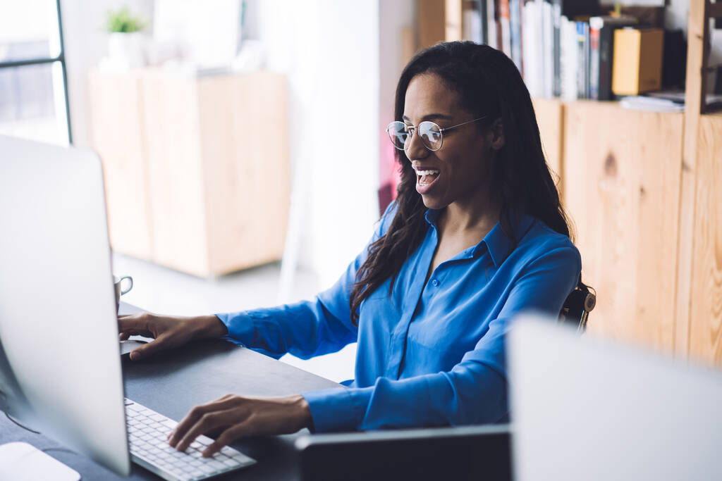 職場の机に座っている正式なシャツの中で魅力的な黒い長い髪の女性と大きなモニターと入力してコンピュータを使用している間、広い笑顔 - 写真・画像