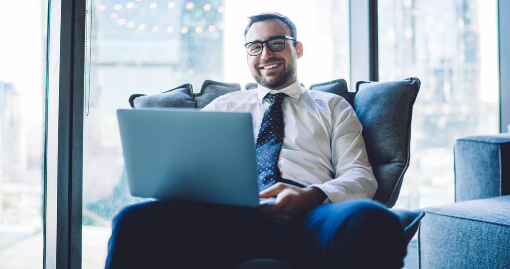 Portret szczęśliwego uśmiechniętego biznesmena siedzącego w biurze i radującego się zakończonym zadaniem na laptopie, wesoły przedsiębiorca w okularach czujący się dobrze z otrzymanych dobrych wiadomości na urządzeniu netbook - Zdjęcie, obraz