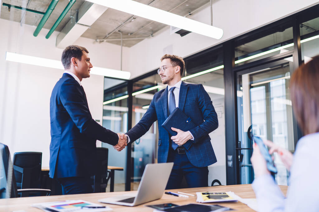 Два уверенных бизнесмена в классической формальной одежде в современном офисе заканчивают встречу и пожимают друг другу руки, глядя друг на друга - Фото, изображение