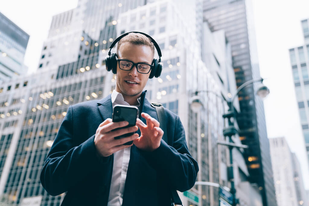 Знизу офіційний красивий чоловік-працівник в окулярах в костюмі насолоджується музикою з бездротовими навушниками, переглядаючи мобільний телефон на розмитому тлі комерційних будівель
  - Фото, зображення