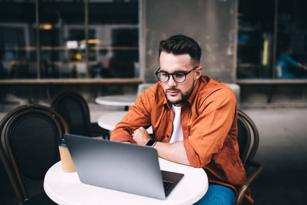 Thoughtful young male freelancer in glasses wearing casual clothes attentively browsing laptop with interest while sitting at table in cafe - Photo, Image