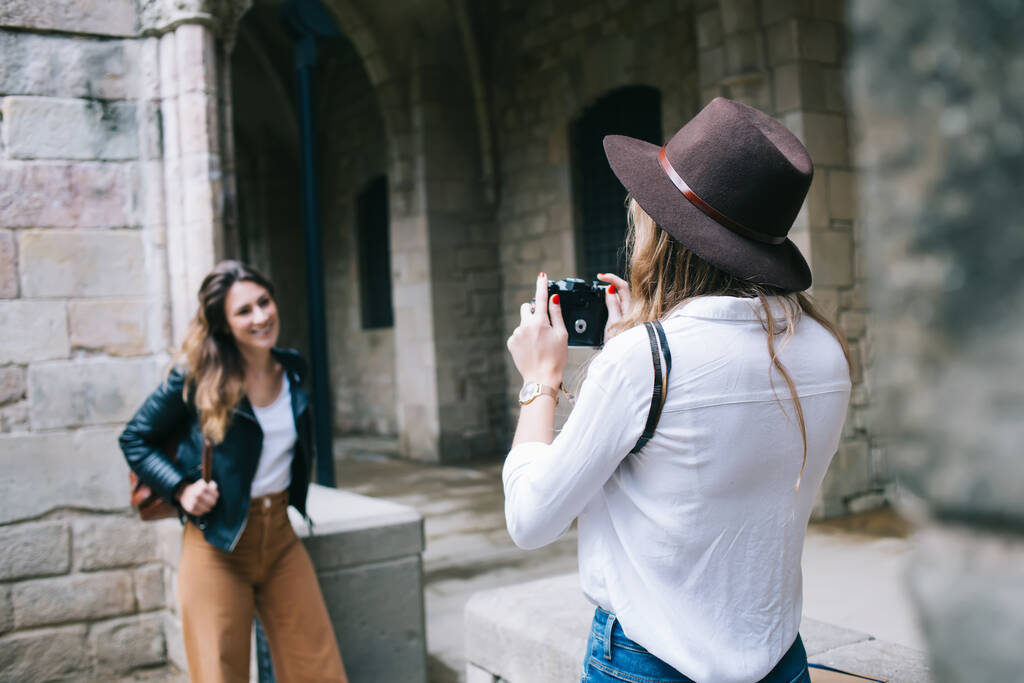 Вид ззаду на невпізнавану жінку в білій сорочці і стильний капелюх стоїть і робить фотографію жінки посміхається компаньйон на кам'яній будівлі
 - Фото, зображення