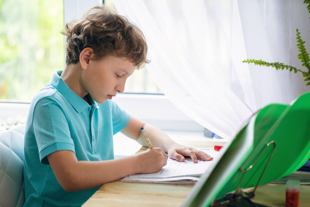 Figyelmes fiú, aki jegyzetfüzetbe ír, miközben az asztalnál ül és házi feladatot csinál. A gyerek házi feladatot csinál otthon. A gyerekek tanulnak és tanulnak. Óvodás gyerek ír és olvas. Vissza az iskolába.. - Fotó, kép