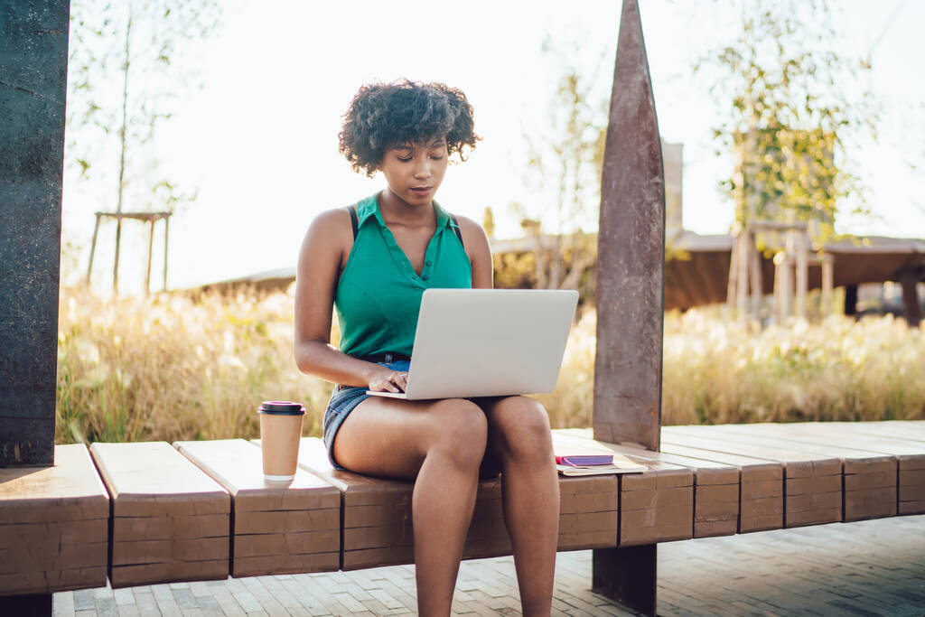 Atractiva mujer de piel oscura concentrada en escribir texto en una computadora portátil trabajando remotamente al aire libre, la bloguera afroamericana creativa comparte contenido multimedia sentado en la calle con una computadora portátil
 - Foto, imagen