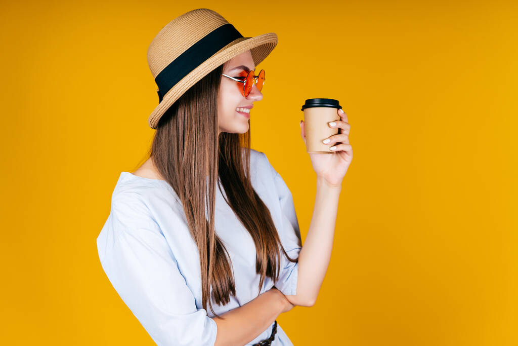 Stilvolles Mädchen mit Hut und Brille hält Kaffee in der Hand und lächelt und schaut irgendwohin. Hintergrund gelb - Foto, Bild