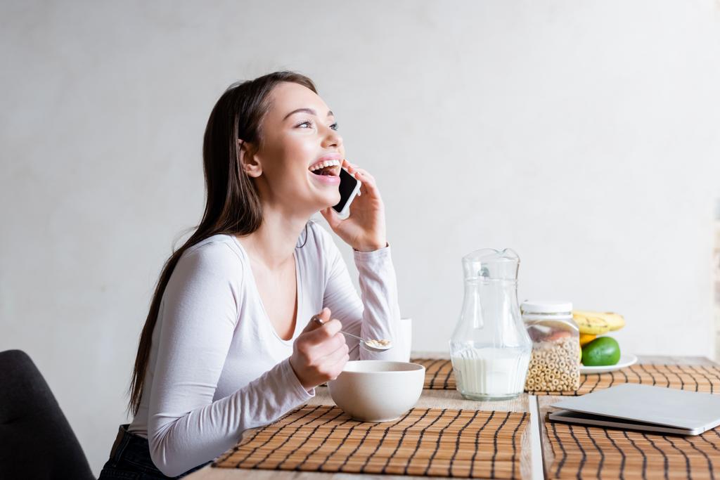 chica feliz hablando en el teléfono inteligente y riendo mientras sostiene la cuchara con sabrosos copos de maíz
  - Foto, Imagen