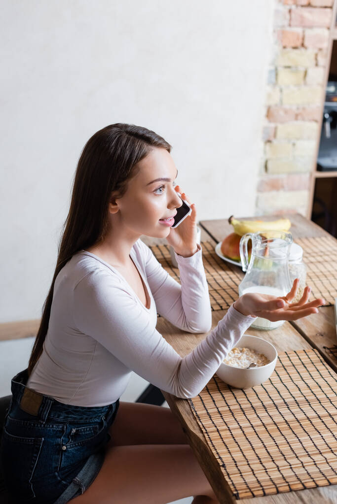 attraktives Mädchen spricht auf Smartphone und gestikuliert in der Nähe von leckeren Cornflakes in der Schüssel - Foto, Bild
