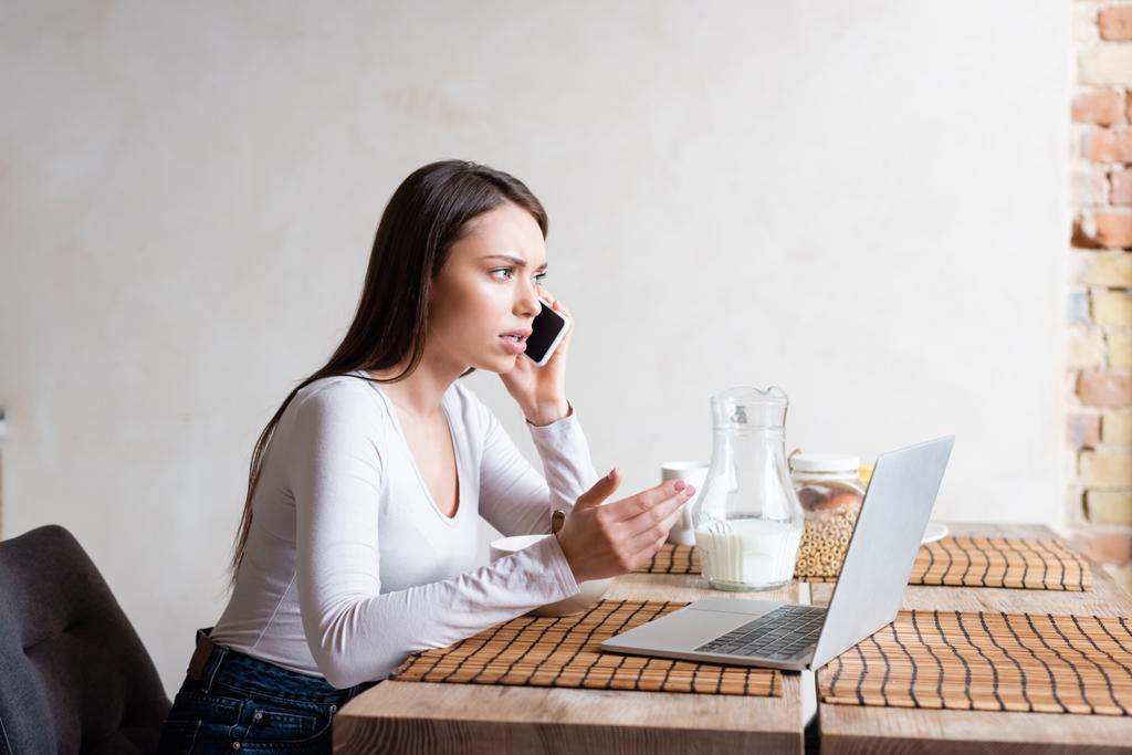 zdenerwowany freelancer rozmowy na smartfonie w pobliżu laptopa i dzbanek z mlekiem  - Zdjęcie, obraz