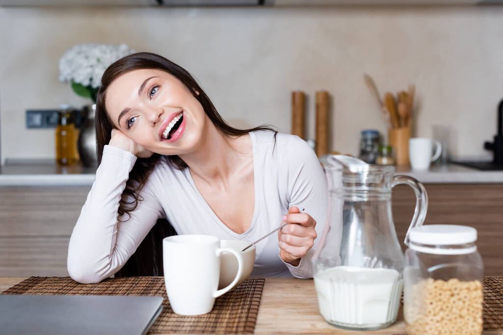 alegre chica sosteniendo cuchara cerca de cuenco, taza y jarra con leche
  - Foto, imagen