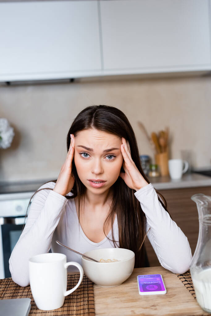 upset girl touching head near bowl, jug and smartphone with online shopping app  - Photo, Image