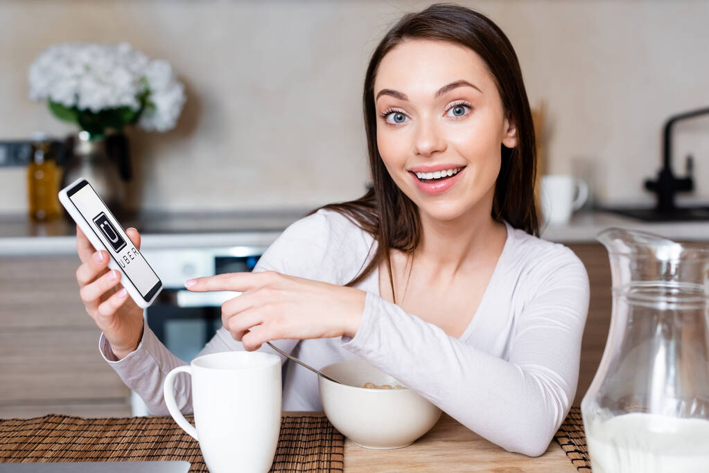 KYIV, UKRAINE - APRIL 29, 2020: вибірковий фокус щасливої дівчини, що вказує пальцем на смартфон з убер-додатком біля чашки, чаші і глечика з молоком.  - Фото, зображення