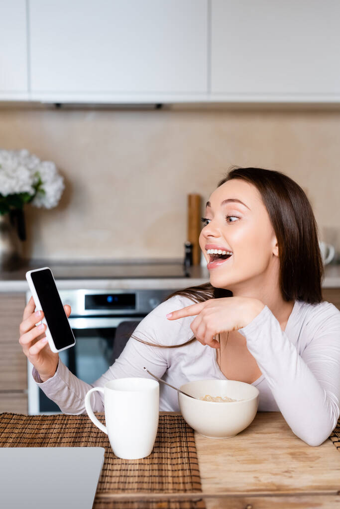 excited girl pointing with finger at smartphone with blank screen near cup and bowl  - Photo, Image
