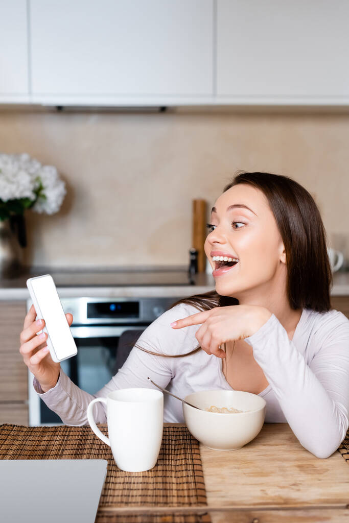 izgatott lány ujjal mutogató okostelefon fehér képernyő közel csésze és tál  - Fotó, kép