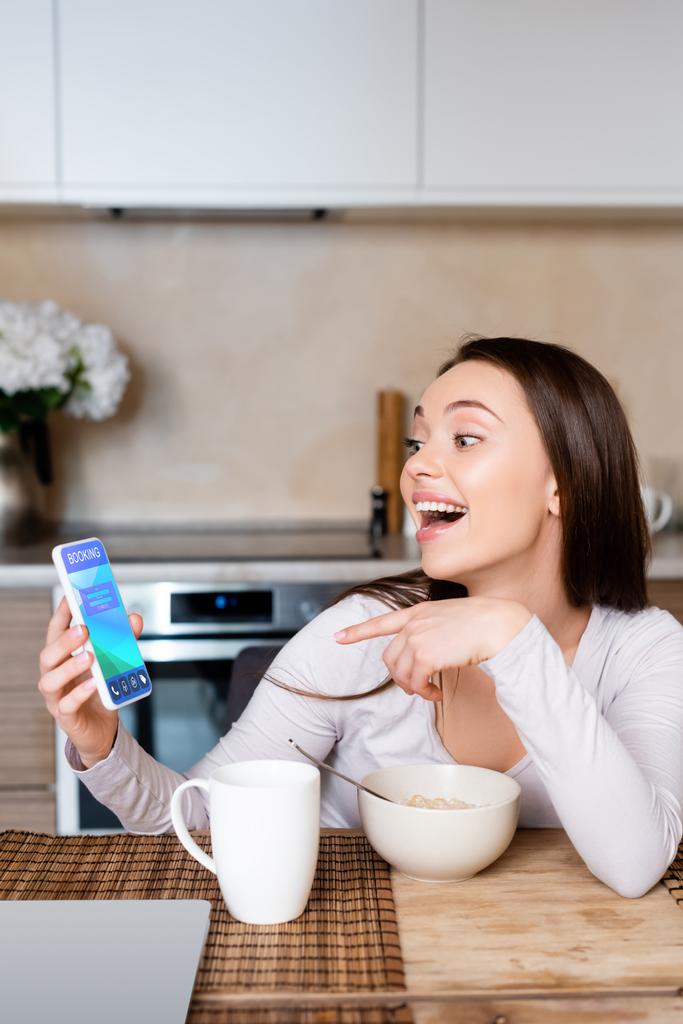 興奮した女性はカップとボウルの近くの予約アプリでスマートフォンを指差す  - 写真・画像