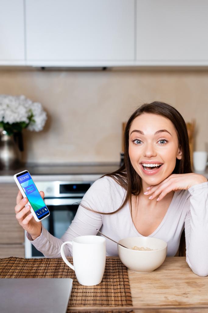 vzrušená žena držící smartphone s rezervační aplikací v blízkosti poháru a mísy  - Fotografie, Obrázek