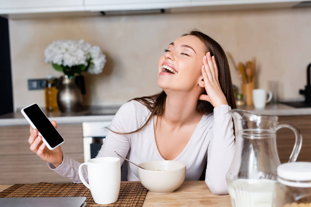 mujer feliz sosteniendo teléfono inteligente con pantalla en blanco y riendo cerca del desayuno
  - Foto, imagen