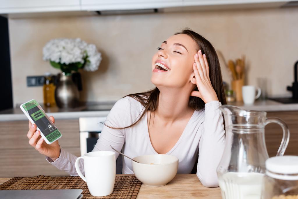 mujer feliz sosteniendo teléfono inteligente con aplicación de reserva y riendo cerca del desayuno
 - Foto, imagen