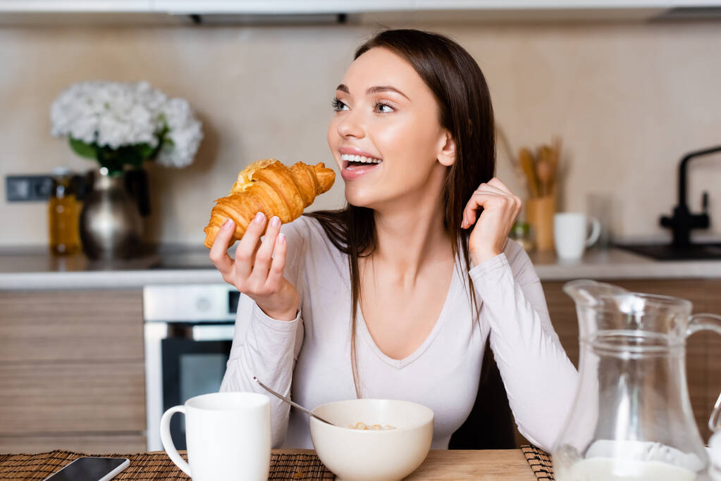 selectieve focus van vrolijk meisje houden croissant in de buurt van smartphone met leeg scherm - Foto, afbeelding