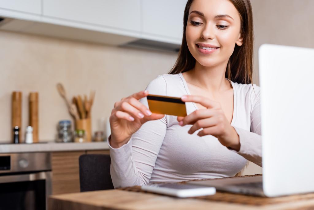 selective focus of cheerful girl holding credit card near laptop and smartphone on table  - Photo, Image
