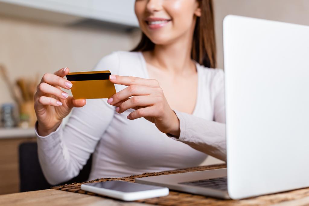 przycięty widok szczęśliwej dziewczyny trzyma kartę kredytową w pobliżu laptopa i smartfona na stole  - Zdjęcie, obraz