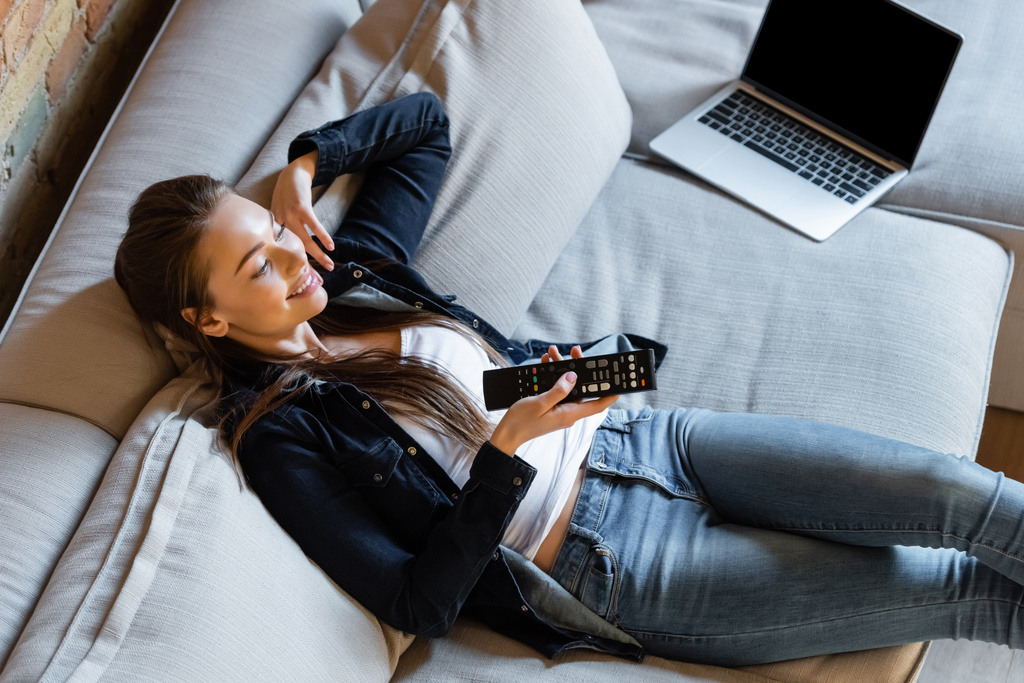 幸せな女性がノートパソコンの近くにリモコンを持っていてソファの上に空白の画面  - 写真・画像