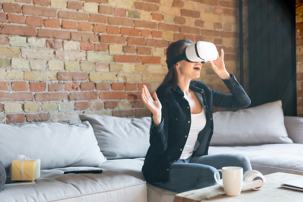 aufgeregte Frau berührt Virtual-Reality-Headset im Sitzen auf dem Sofa - Foto, Bild