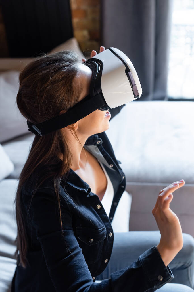 junge Frau gestikuliert mit Virtual-Reality-Headset im Wohnzimmer - Foto, Bild